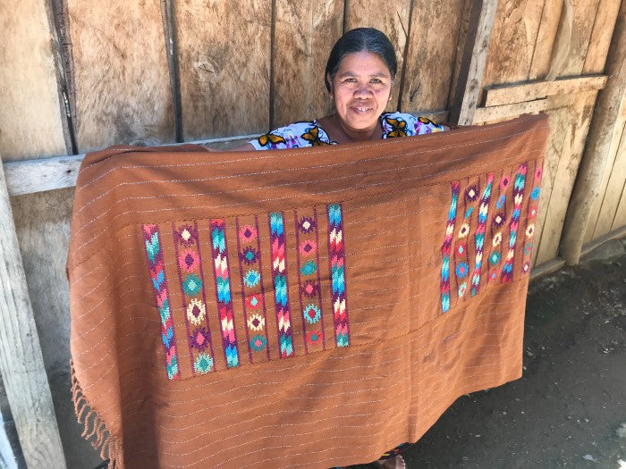 Woman holding a large brown fabric with colorful patterns in front of a wooden wall.