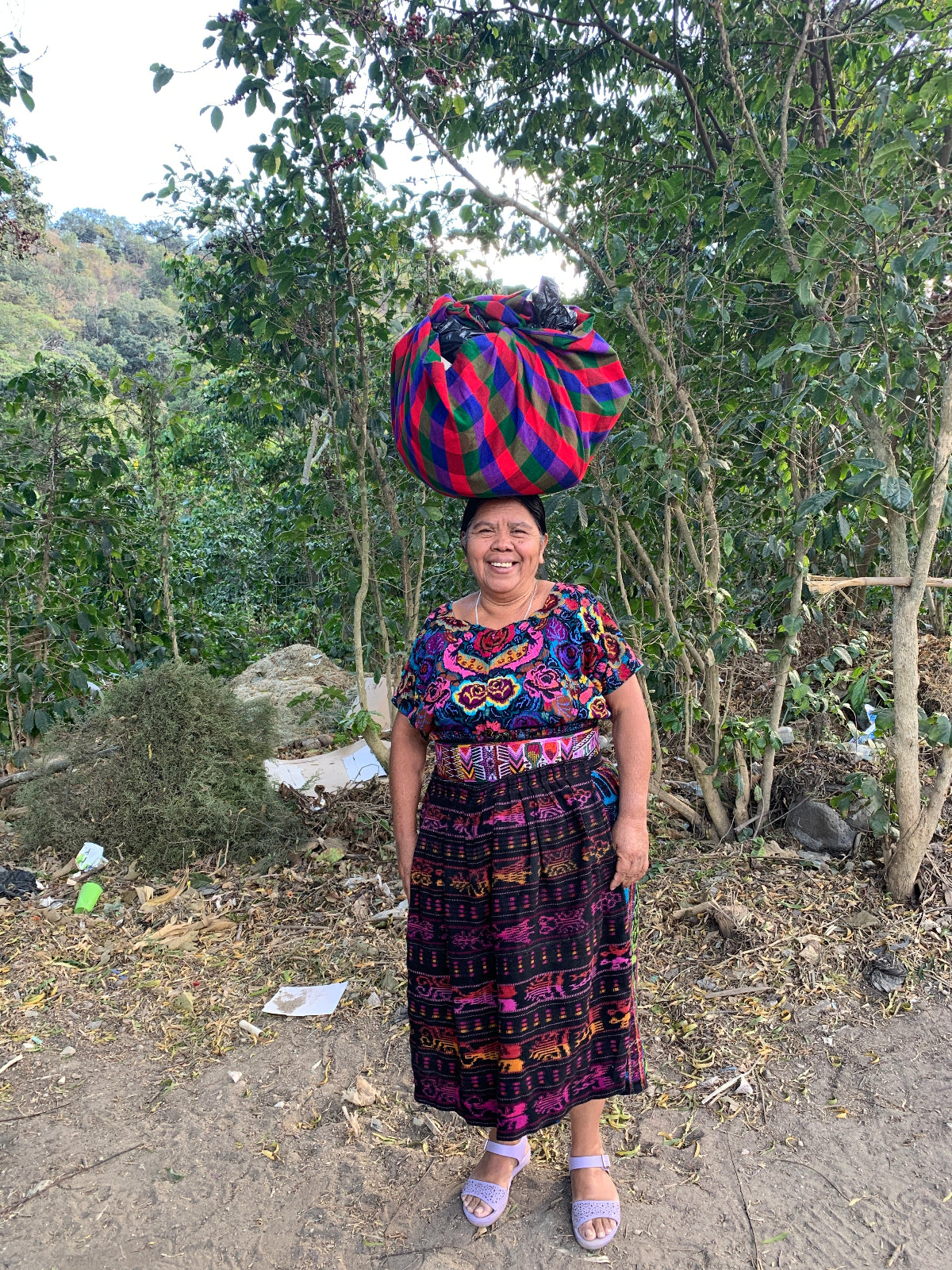 Woman in traditional colorful huipil, corte, apron, and sash standing outdoors carrying a bundle on her head