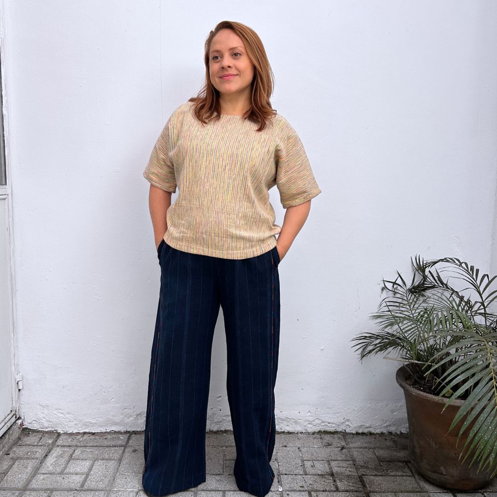 Woman wearing a beige top and dark pants standing against a white wall with a plant to her right.