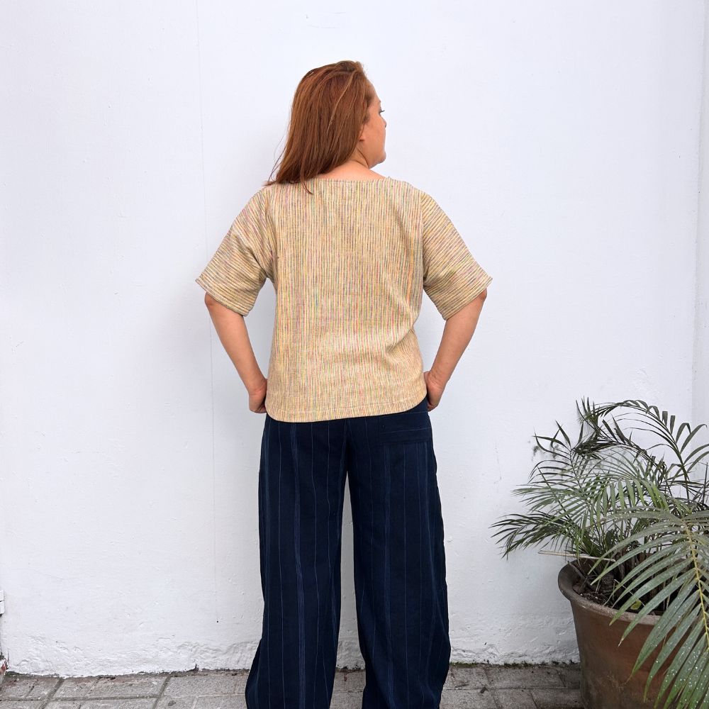 Model Striped multicolor top on a  and dark blue pants standing against a white wall with a plant to the right.
