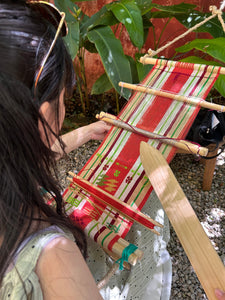 Person using a small wooden loom with striped warp, surrounded by plants.