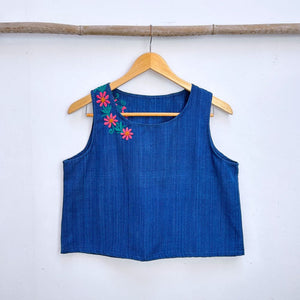 Blue sleeveless top with floral embroidery on a wooden hanger against a white wall.