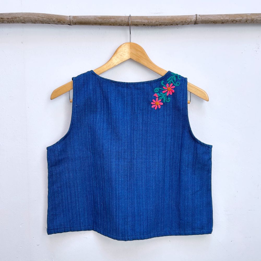 Blue sleeveless top with floral embroidery on a wooden hanger against a white wall.