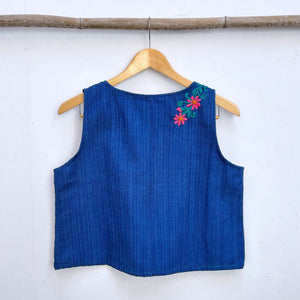 Blue sleeveless top with floral embroidery on a wooden hanger against a white wall.
