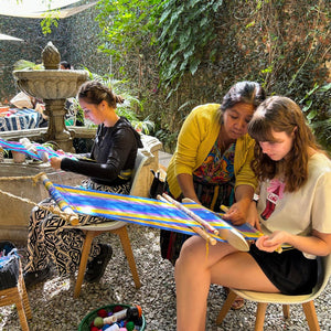 A master weaver teaching two students traditional backstrap loom weaving techniques.