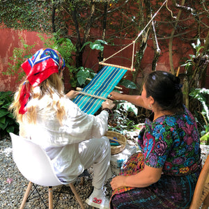Two people engaged in backstrap weaving at a studio garden, with one person wearing a white outfit and the other in colorful traditonal wear.