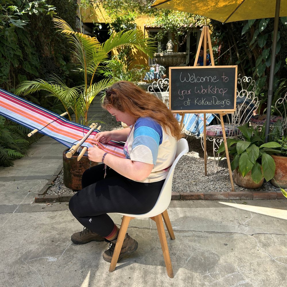 A person seated in the Kakaw Designs garden with a colorful backstrap loom, surrounded by plants and a welcome sign.