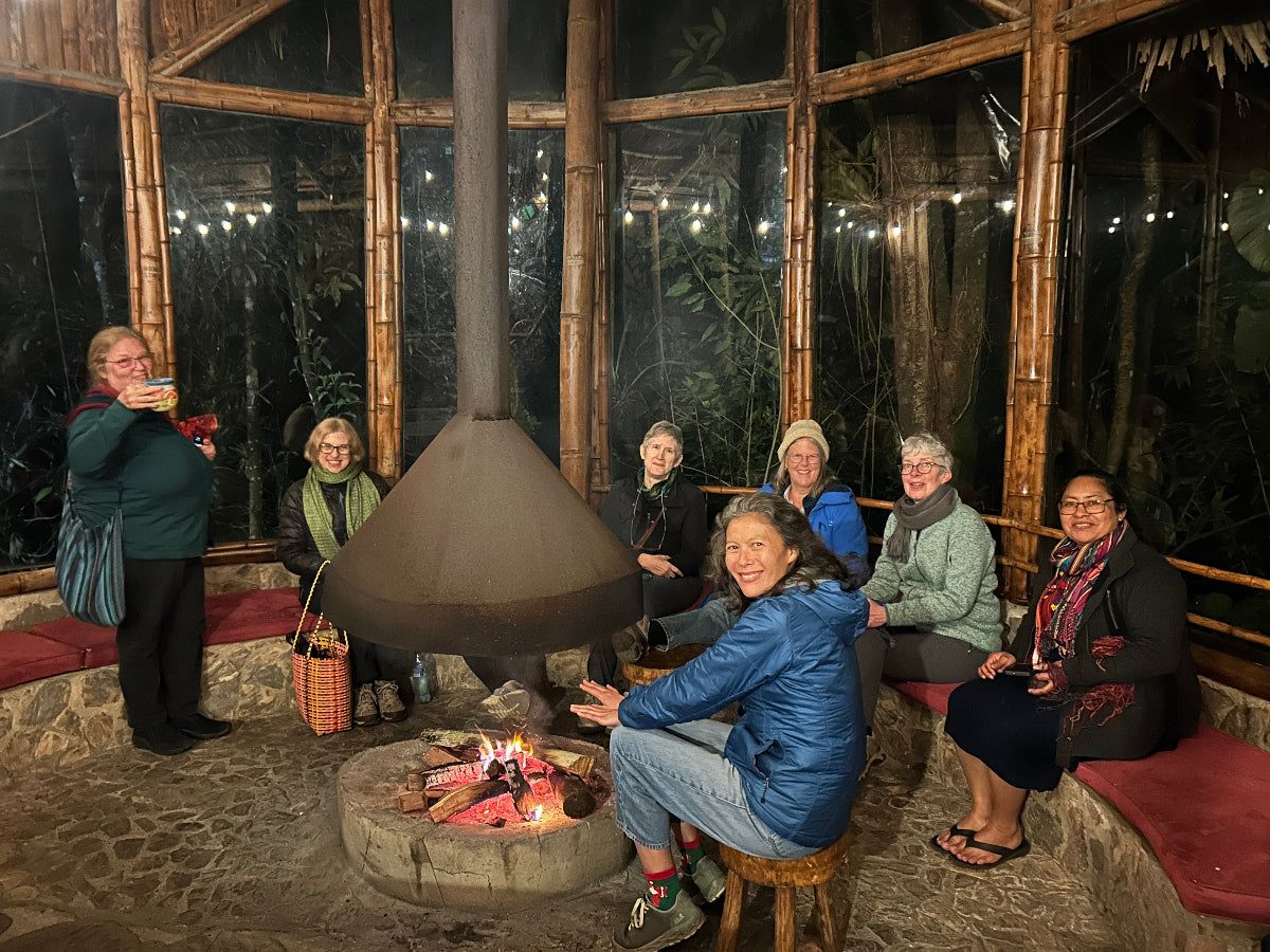 Group of people gathered around a fire pit in a rustic setting with large windows.