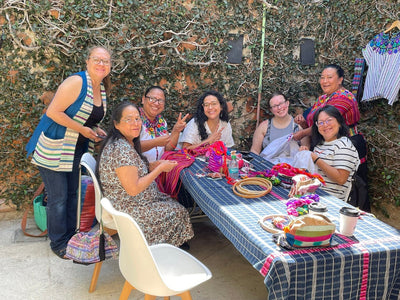 Group of women gathered around a table with colorful textiles and threads, outdoors.