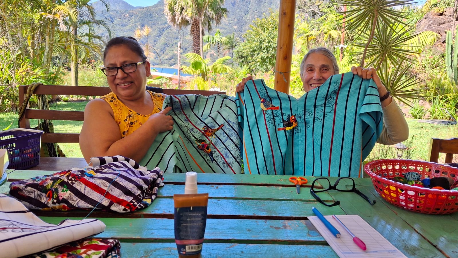 Two women sitting at a table outdoors, holding up the huipiles they are embroidering