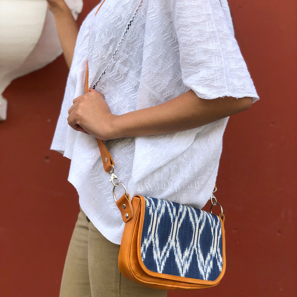 A person wearing a white shirt is holding a clutch bag with a geometric ikat pattern in blue and white, and a tan leather strap and back compartment.