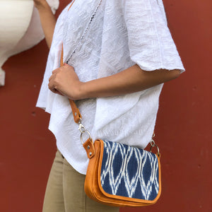 A person wearing a white shirt is holding a clutch bag with a geometric ikat pattern in blue and white, and a tan leather strap and back compartment.