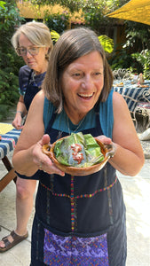 A woman in a blue apron smiles while holding a small wooden bowl filled with banana leaves, tamalitos and a red sauce, set in an outdoor patio environment with another woman in the background.