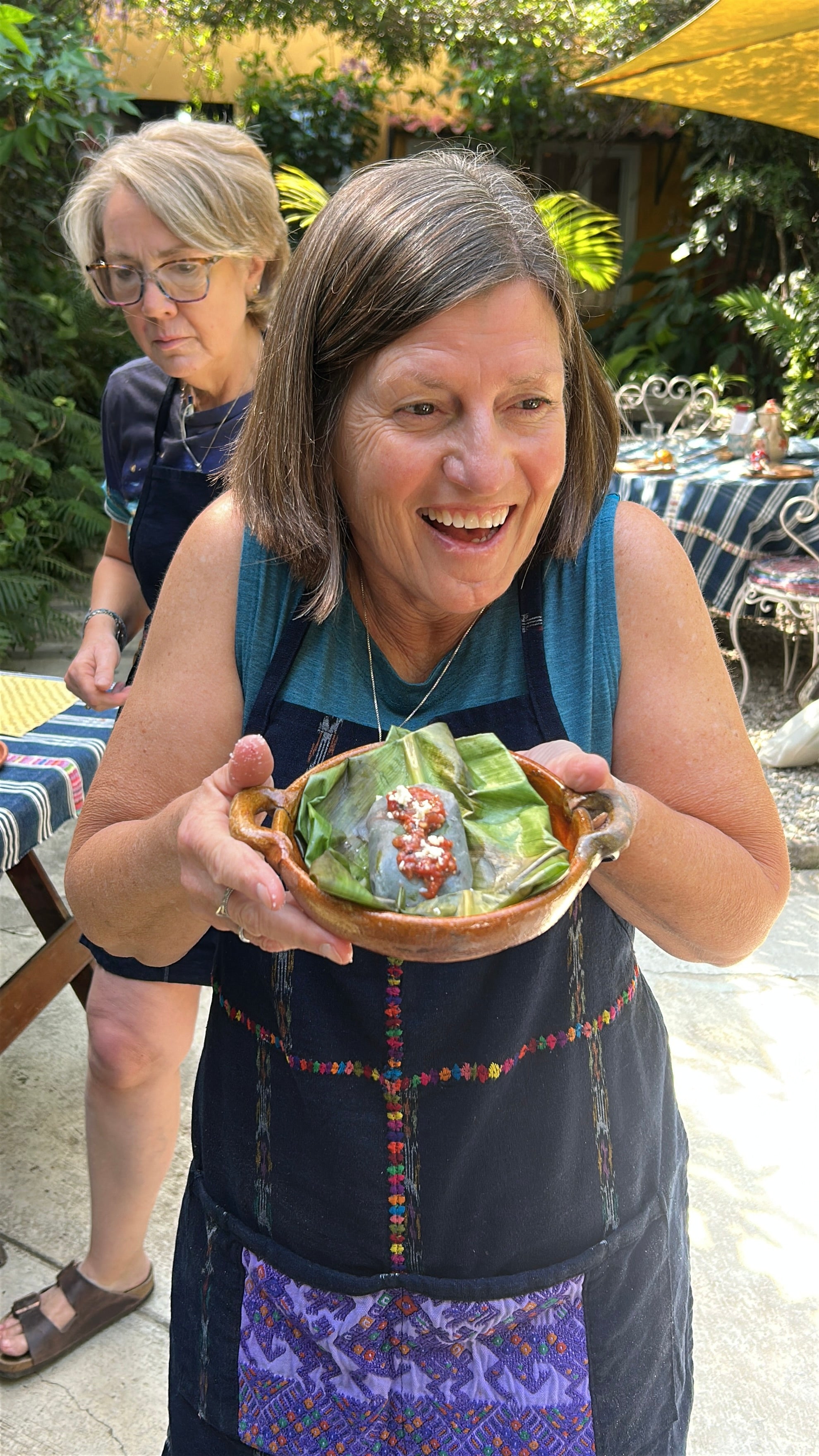 A woman in a blue apron smiles while holding a small wooden bowl filled with banana leaves, tamalitos and a red sauce, set in an outdoor patio environment with another woman in the background.