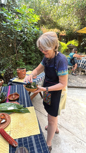 A woman wearing a navy blue apron over a dark dress stands outdoors at a table, holding a small bowl with tamalitos. The table is decorated with woven mats, a corn cob, and other food items, set against a lush garden background.
