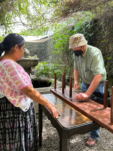Two people standing near a wooden table outdoors with trees in the background