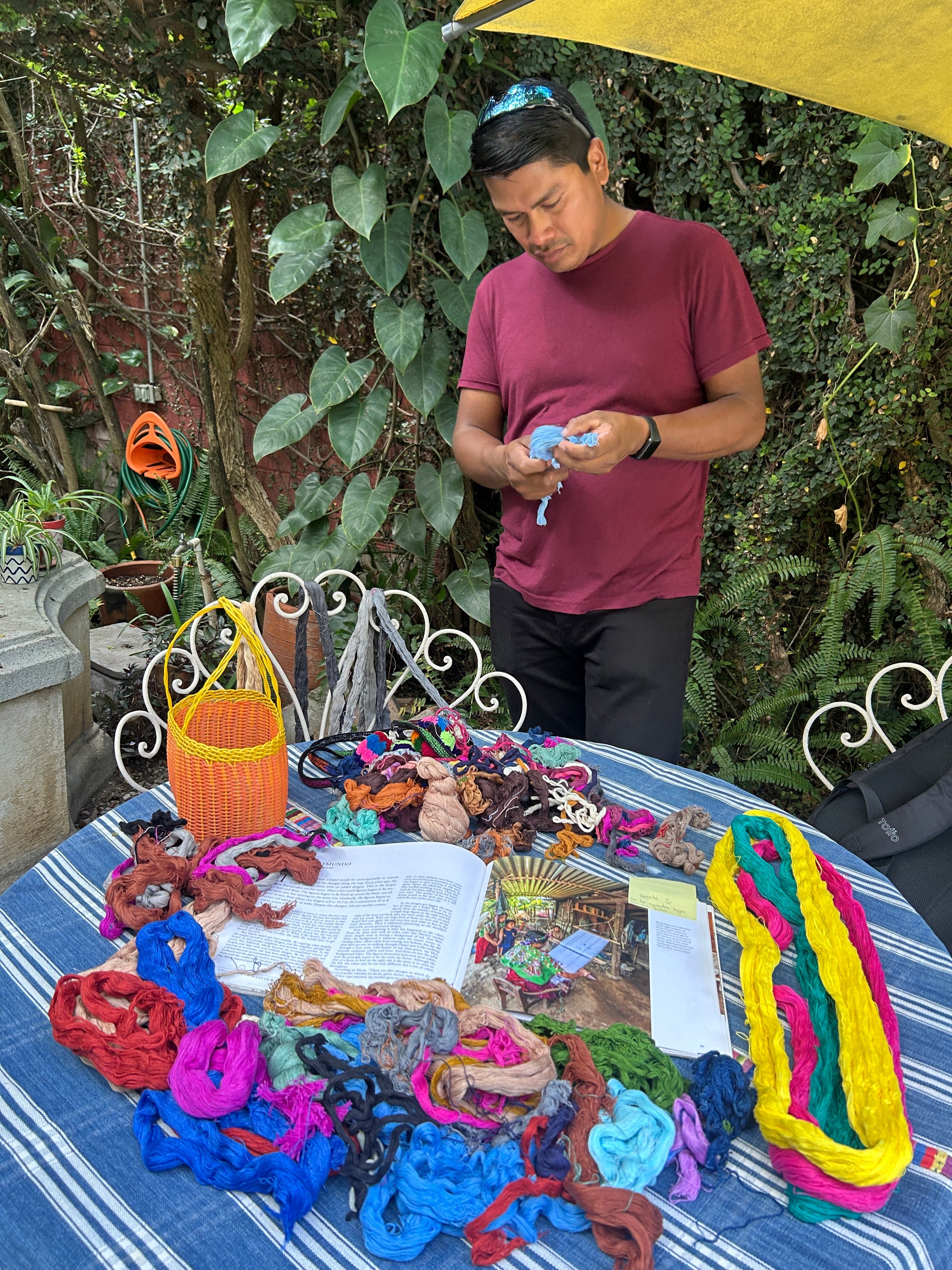 Chato, our master pompom teacher, is seated in the Kakaw garden holding naturally dyed cotton yarns used to create the central structure of a pompom. In front of him is a table with all the materials used during the class, including yarns, scissors, and brushes.
