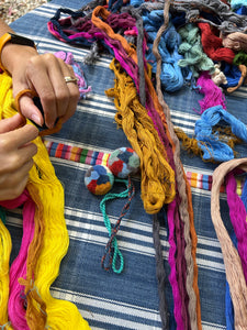A table covered with naturally dyed cotton yarns in a variety of colors. In the center are two finished pompoms featuring vibrant, multicolored designs.