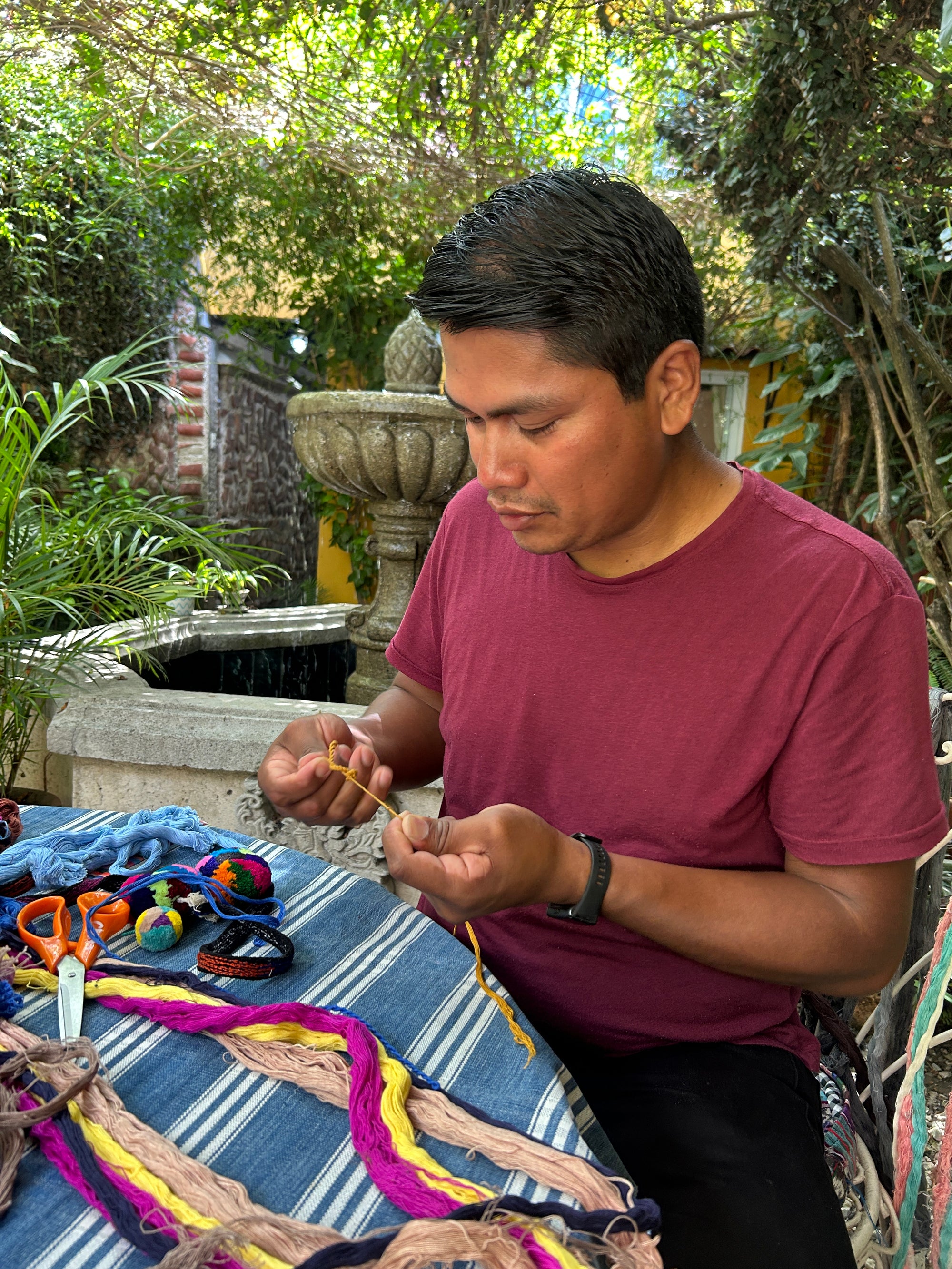 Chato, our master pompom teacher, is seated in the Kakaw garden holding naturally dyed cotton yarns used to create the central structure of a pompom. In front of him is a table with all the materials used during the class, including yarns, scissors, and brushes.