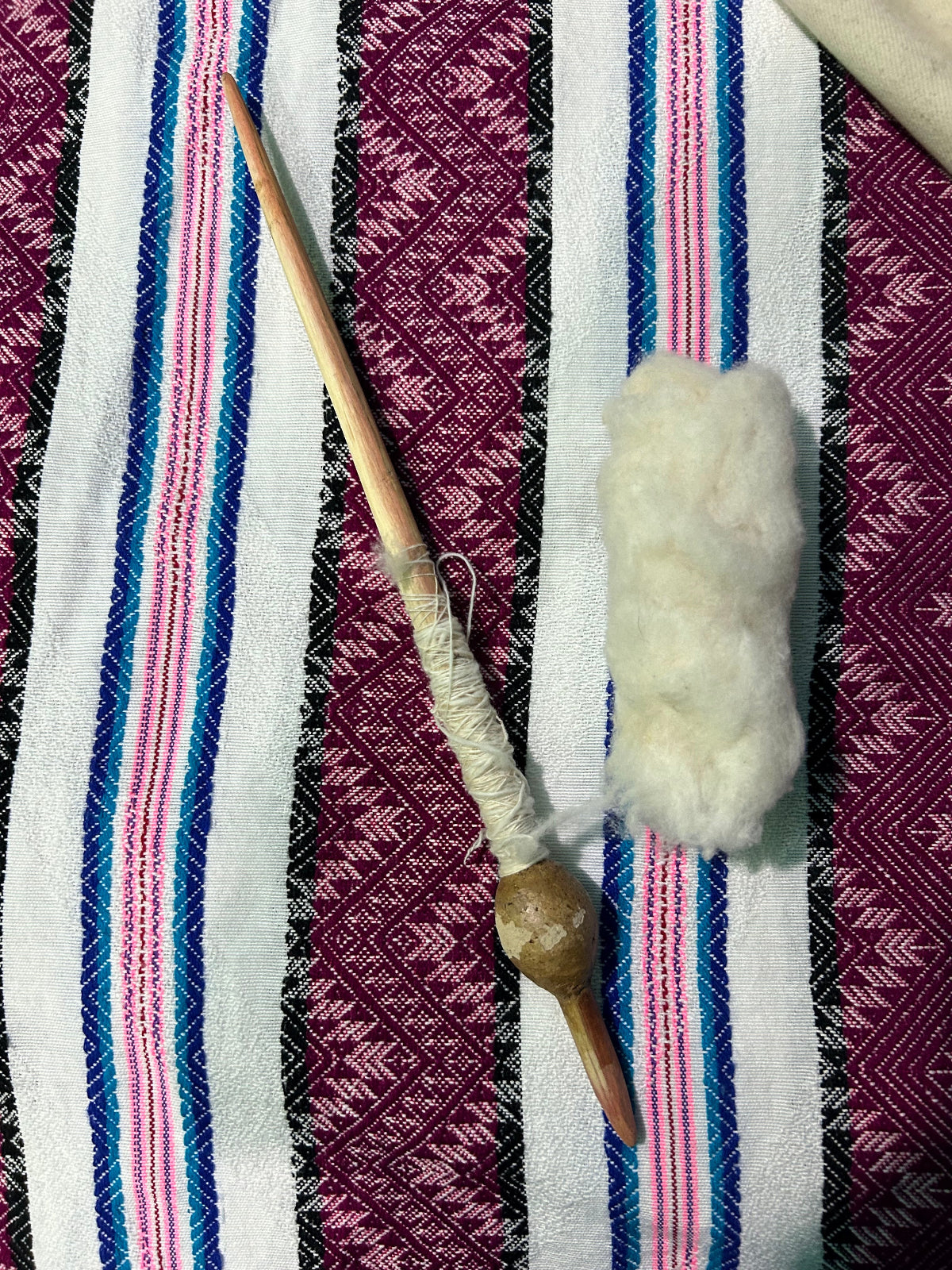 A traditional wooden spinning tool with a wooden bobbin and a bundle of white cotton fiber, laid out on a colorful woven textile background.