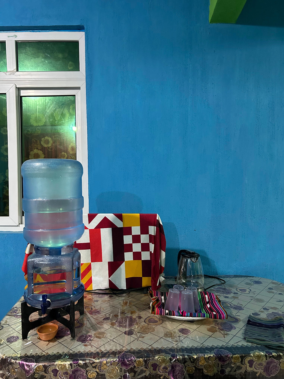A large plastic water dispenser bottle filled with filter water sits on a small black stand on a patterned tablecloth, set against a blue wall in an indoor room. A colorful quilted blanket, a glass, and other household items are visible nearby.