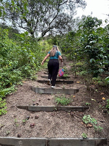 A woman with long hair is seen from behind, walking up a dirt path with concrete steps on a lush, green outdoor path.