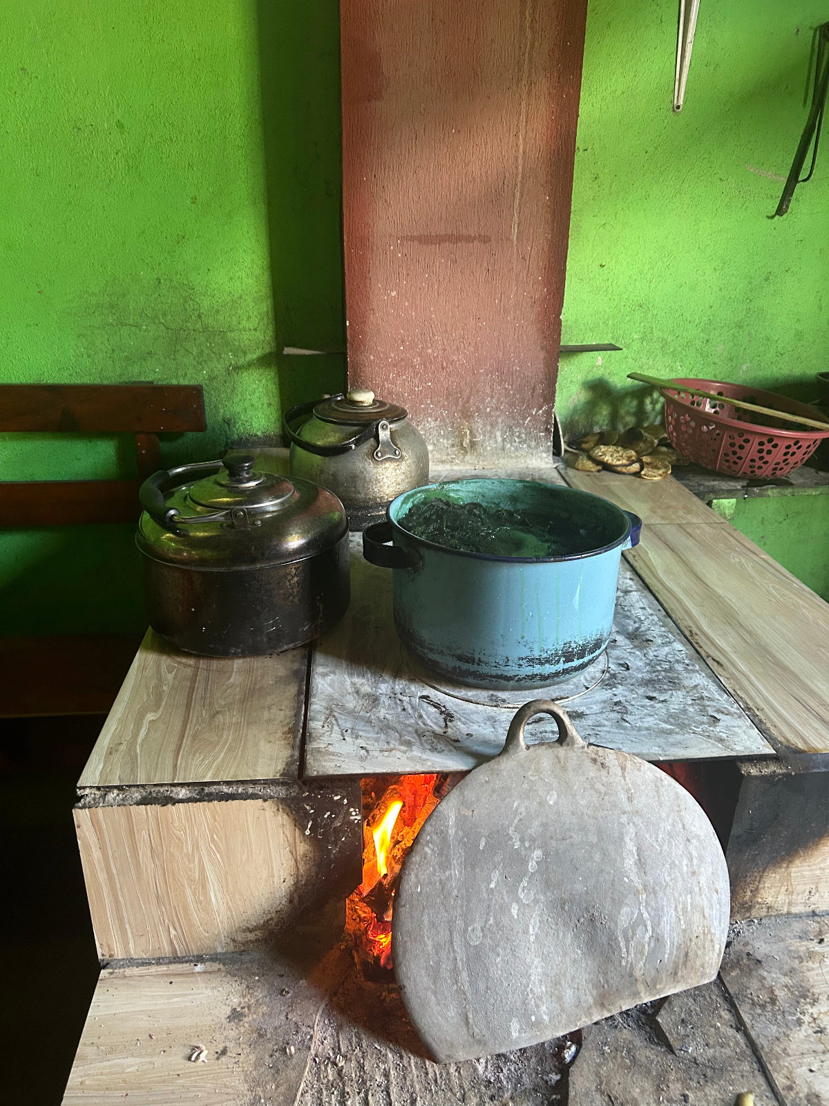 A rustic wood-burning stove with a visible fire, holding several cooking pots and a large stone lid or cover leaning in front. The scene is set against a bright green wall in a real indoor environment.