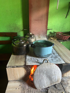 A rustic wood-burning stove with a visible fire, holding several cooking pots and a large stone lid or cover leaning in front. The scene is set against a bright green wall in a real indoor environment.