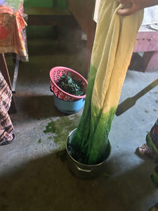 A person holds up a long, narrow fabric that is being dyed green, dipping the lower portion into a metal bucket filled with dark green pigment. A pink plastic basket containing more green pigment sits on a blue bucket on the concrete floor nearby.