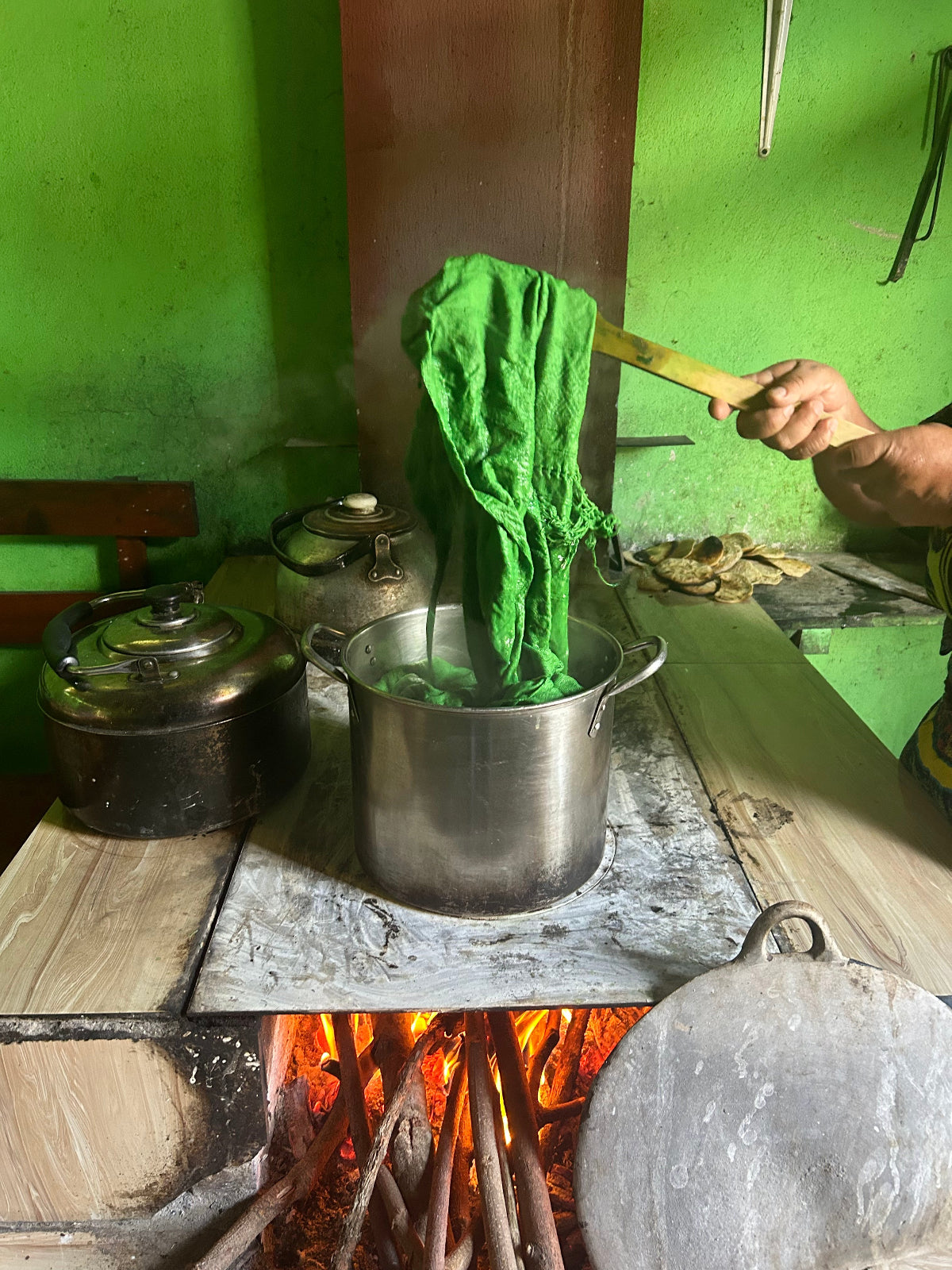 A person's hand holds a green cloth or garment over a metal pot on a rustic wood-burning stove, with other cooking pots and a green wall in the background.