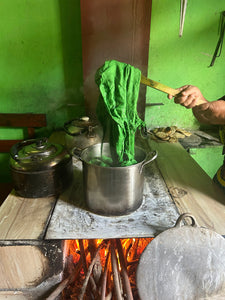 A person's hand holds a green cloth or garment over a metal pot on a rustic wood-burning stove, with other cooking pots and a green wall in the background.