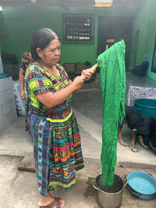 A woman in a colorful Guatemalan-style patterned dress stands indoors holding up a large green cloth or garment over a metal pot on a concrete floor. A dog is visible in the background near a table and tubs.