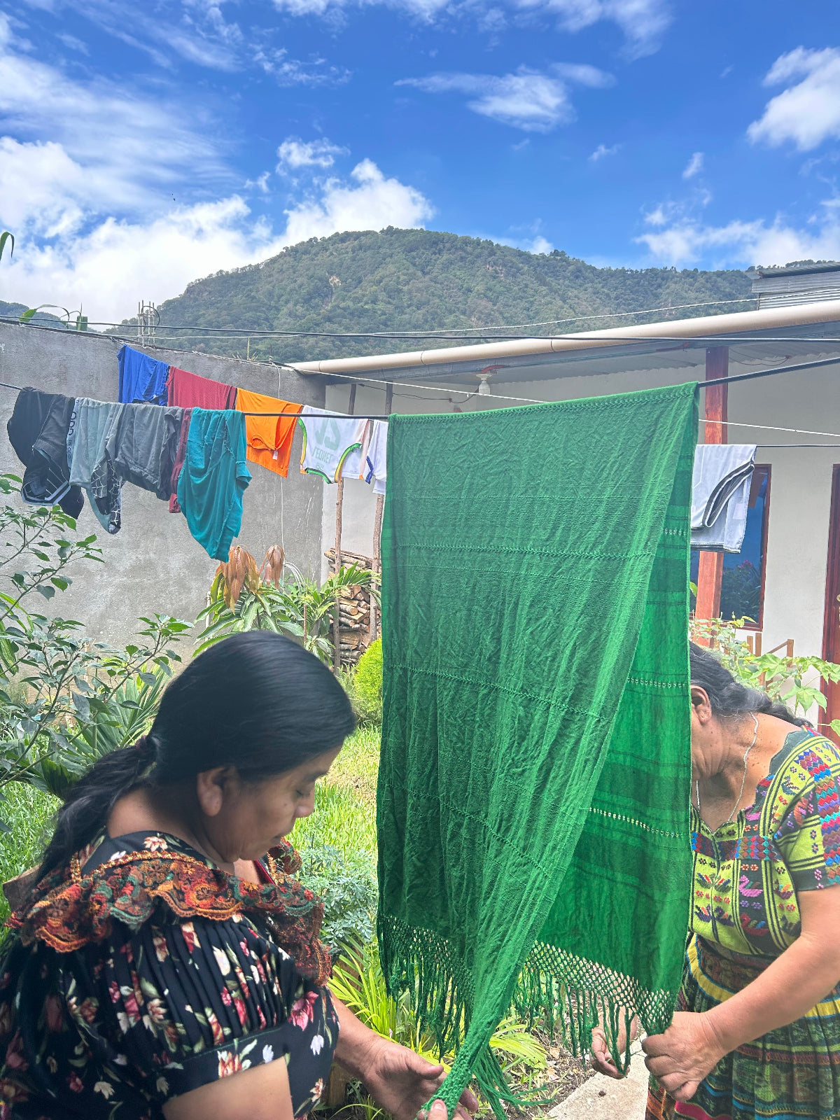 A large green woven textile, likely a blanket or scarf, is being held up outdoors by two women in a backyard setting with laundry hanging on a line and a mountain in the background.