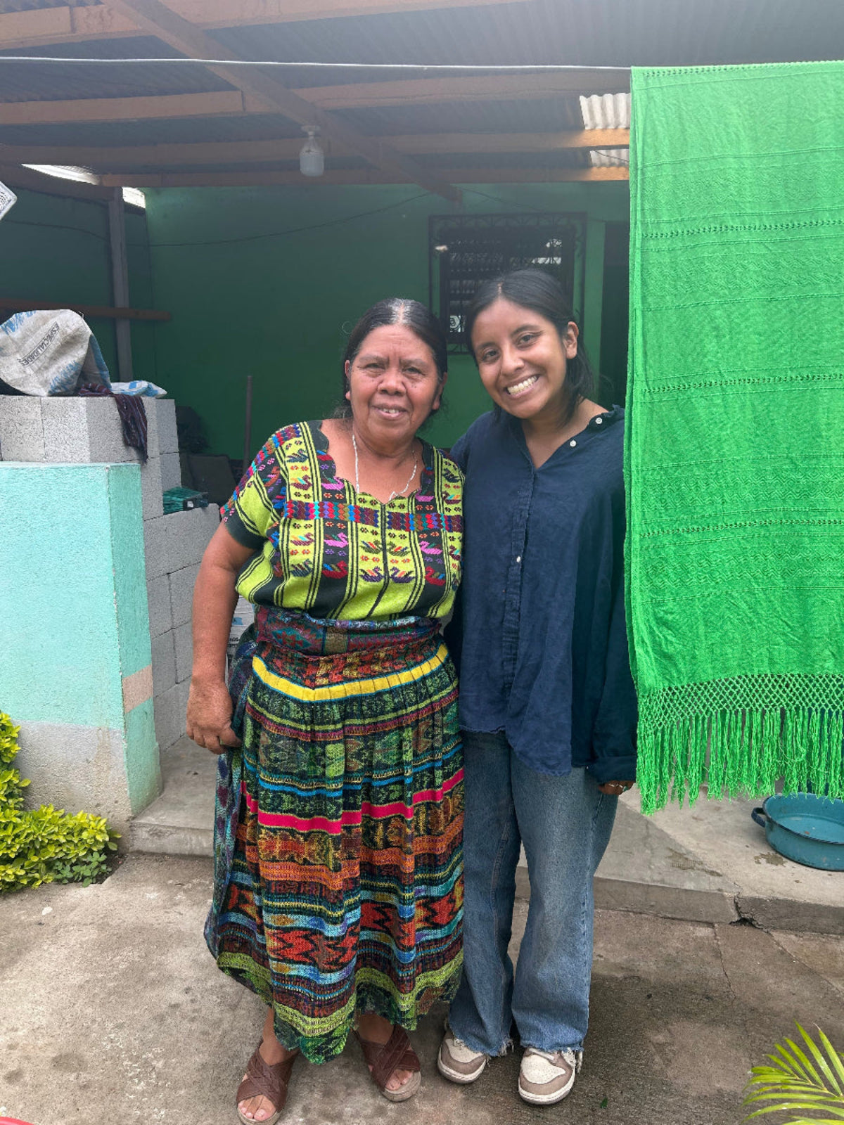 Two women standing together in front of a home entrance. The woman on the left wears a colorful traditional Guatemalan-style weaving blouse and a long striped skirt with green, yellow, red, blue, and black patterns.