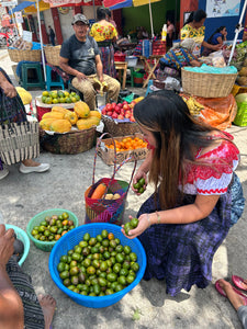 A woman in a red and white embroidered blouse and a dark patterned skirt bends over at a rustic outdoor local market stall, reaching into a blue basket filled with jocotes fruits. Other market vendors and produce are visible in the background.