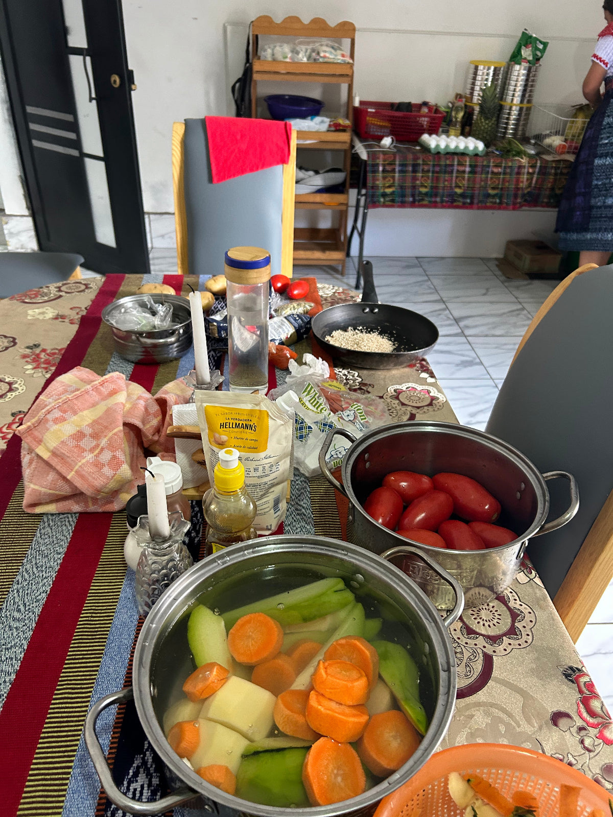 A home kitchen scene showing a stainless steel pot filled with vegetables, including carrots, celery, and potatoes, alongside another pot of tomatoes, on a patterned table runner.