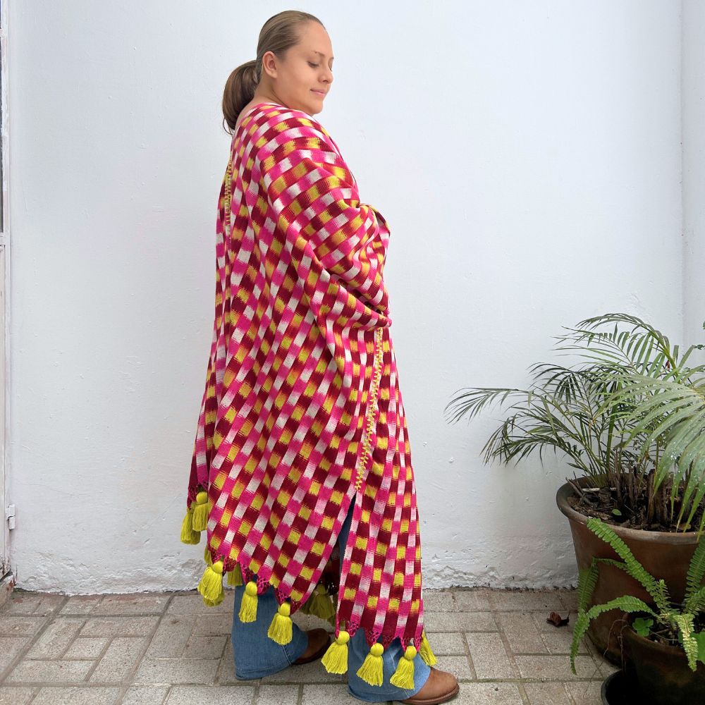 Woman wearing a colorful checkered shawl with tassels against a white wall.
