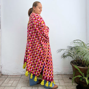 Woman wearing a colorful checkered shawl with tassels against a white wall.