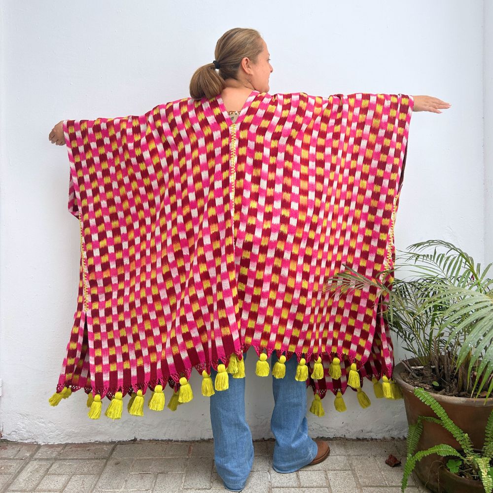 Person wearing a colorful pink and yellow checkered textile garment with tassels, standing against a white wall.