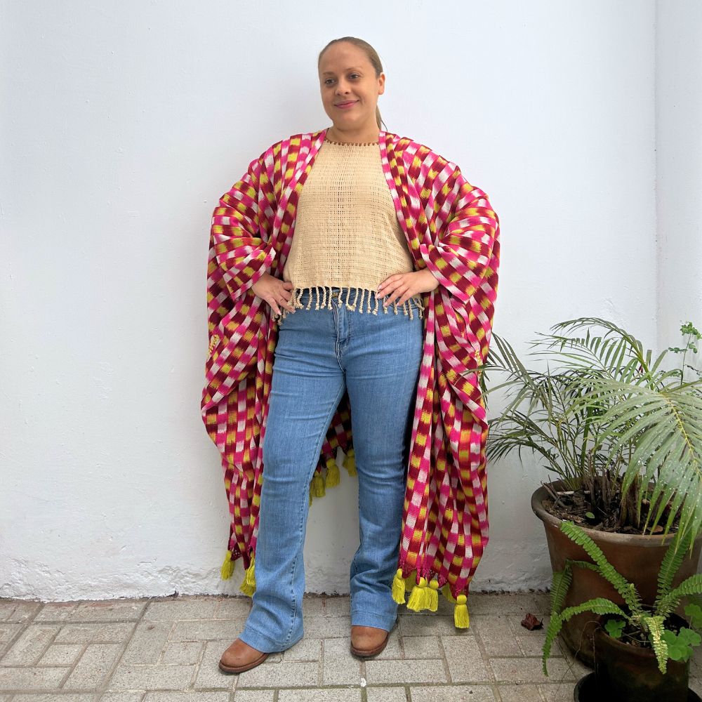 Person wearing a colorful patterned shawl standing next to a plant against a white wall.