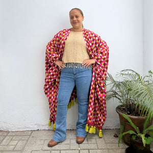 Person wearing a colorful patterned shawl standing next to a plant against a white wall.
