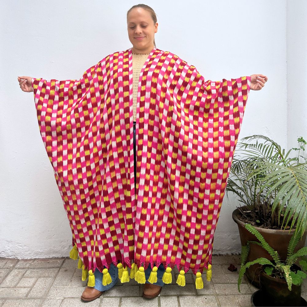 Person wearing a colorful checkered textile garment with tassels, standing next to a plant 