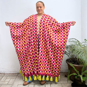 Person wearing a colorful checkered textile garment with tassels, standing next to a plant 