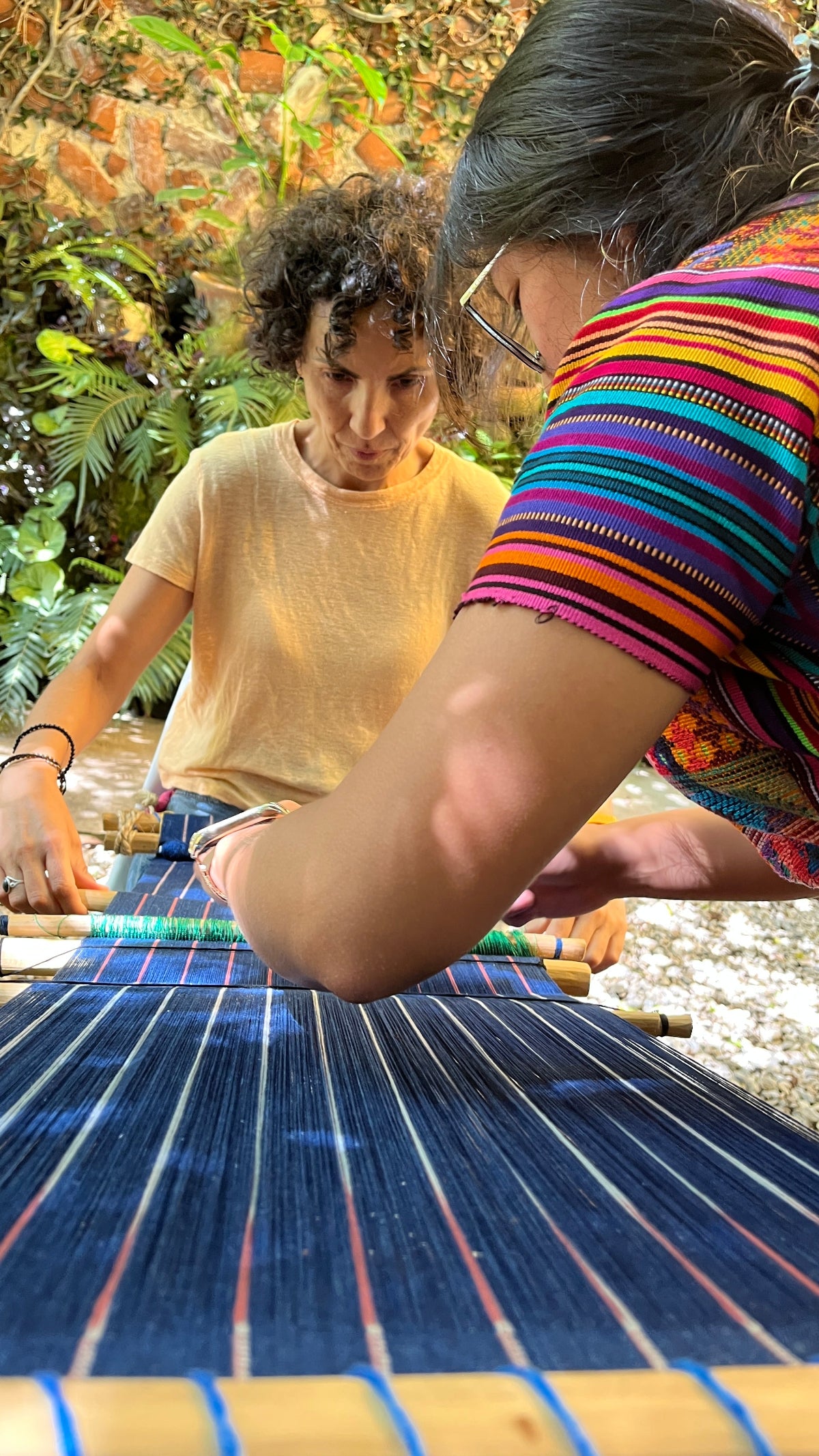 Two people working on an indigo-dyed bacsktrap loom outside
