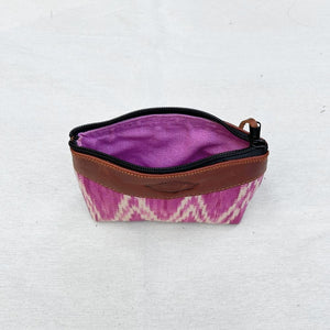 A zippered pouch with a pink and white ikat pattern exterior, brown leather trim, and a purple interior lining, shown on a light fabric background.