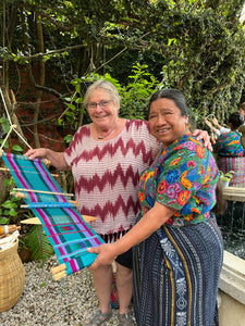 Weaving instructor in traditional clothing holding a colorful woven item outdoors with her student.