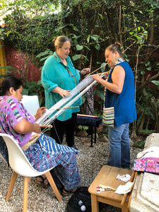 Three women working with a loom outdoors, surrounded by plants and tools.
