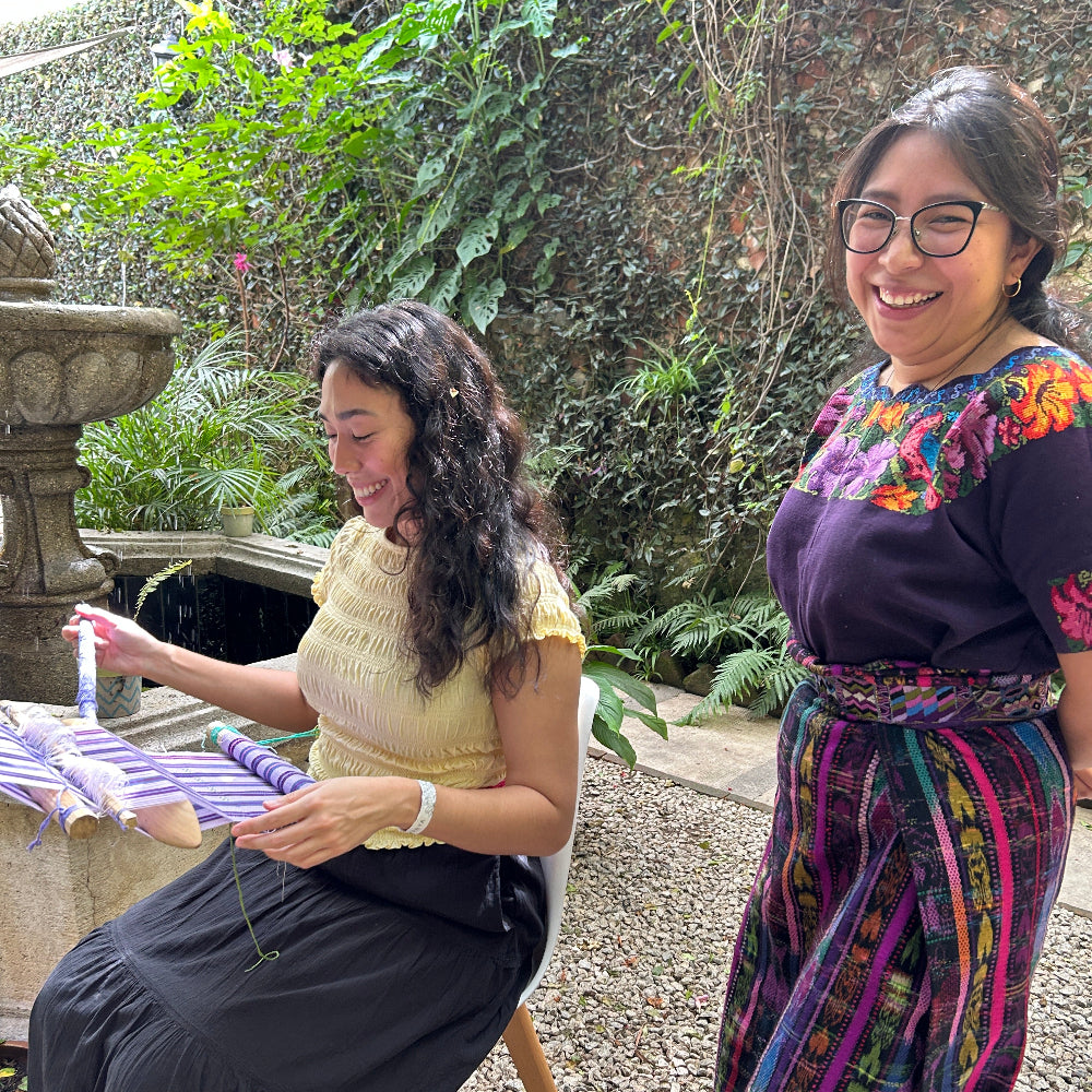 Two women standing outdoors with a stone structure and greenery in the background