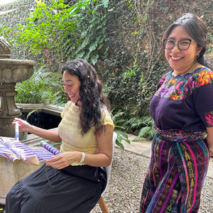 Two women standing outdoors with a stone structure and greenery in the background