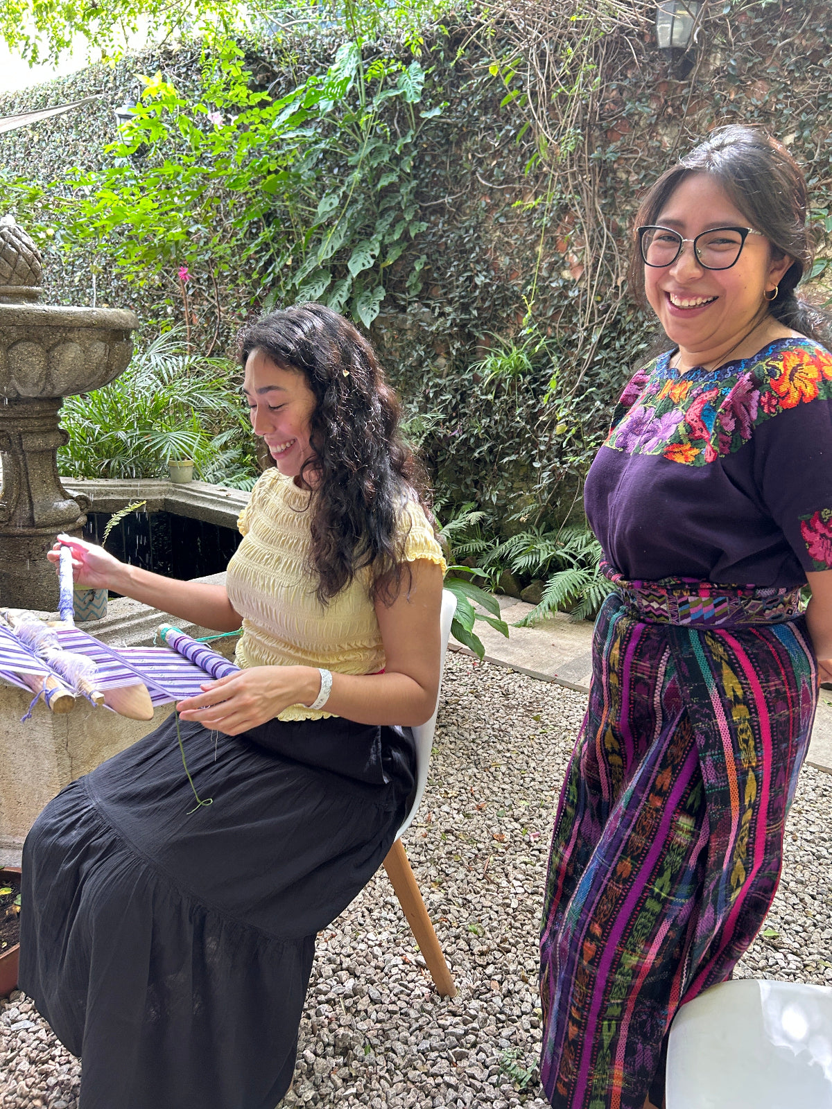 Student weaving on a backstrap loom, and teacher observing, both with a big smile.
